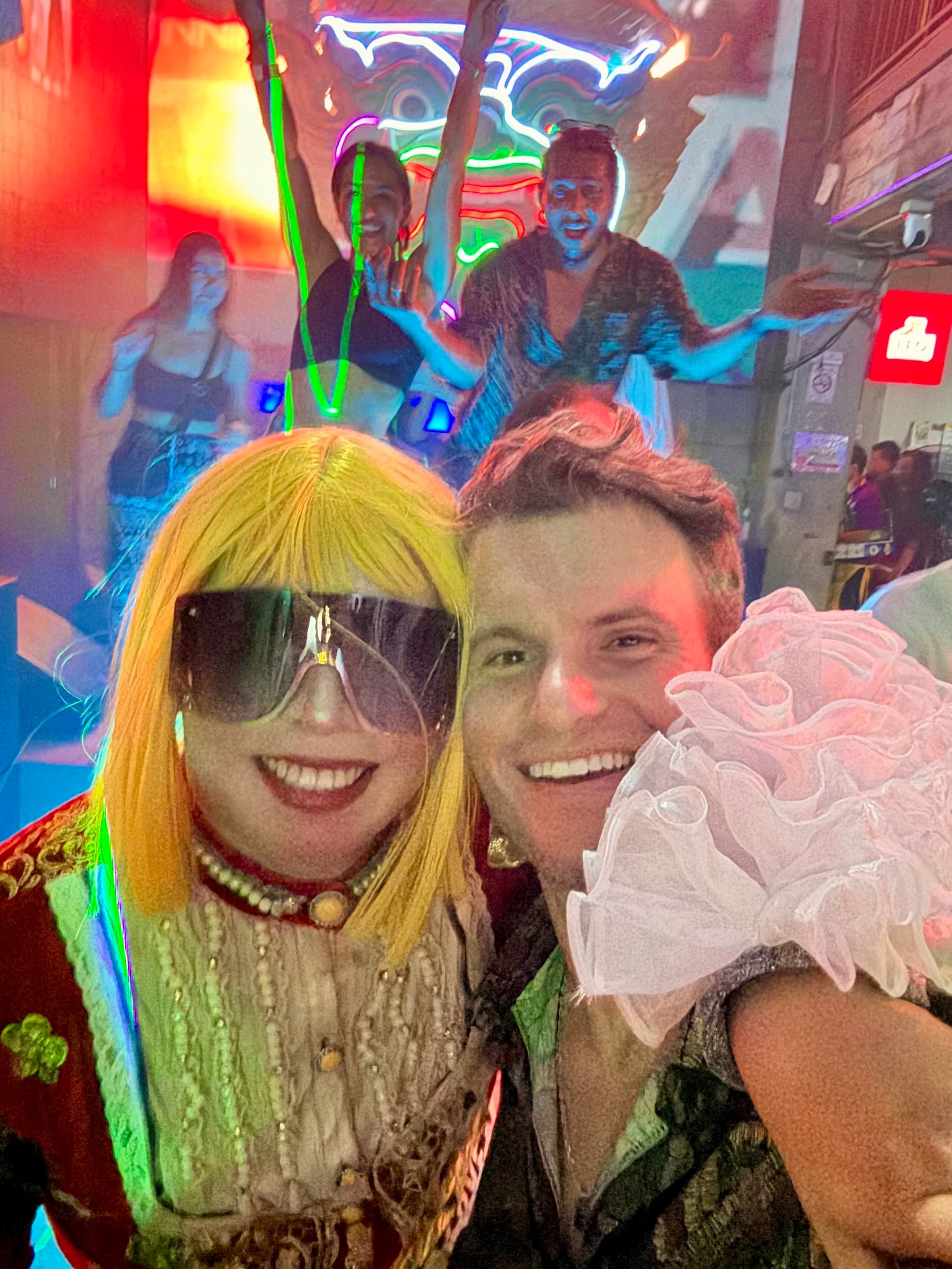 A traveler taking a nighttime selfie with a costumed cabaret performer in a blonde wig and oversized sunglasses under neon lights on a Bangkok nightlife street