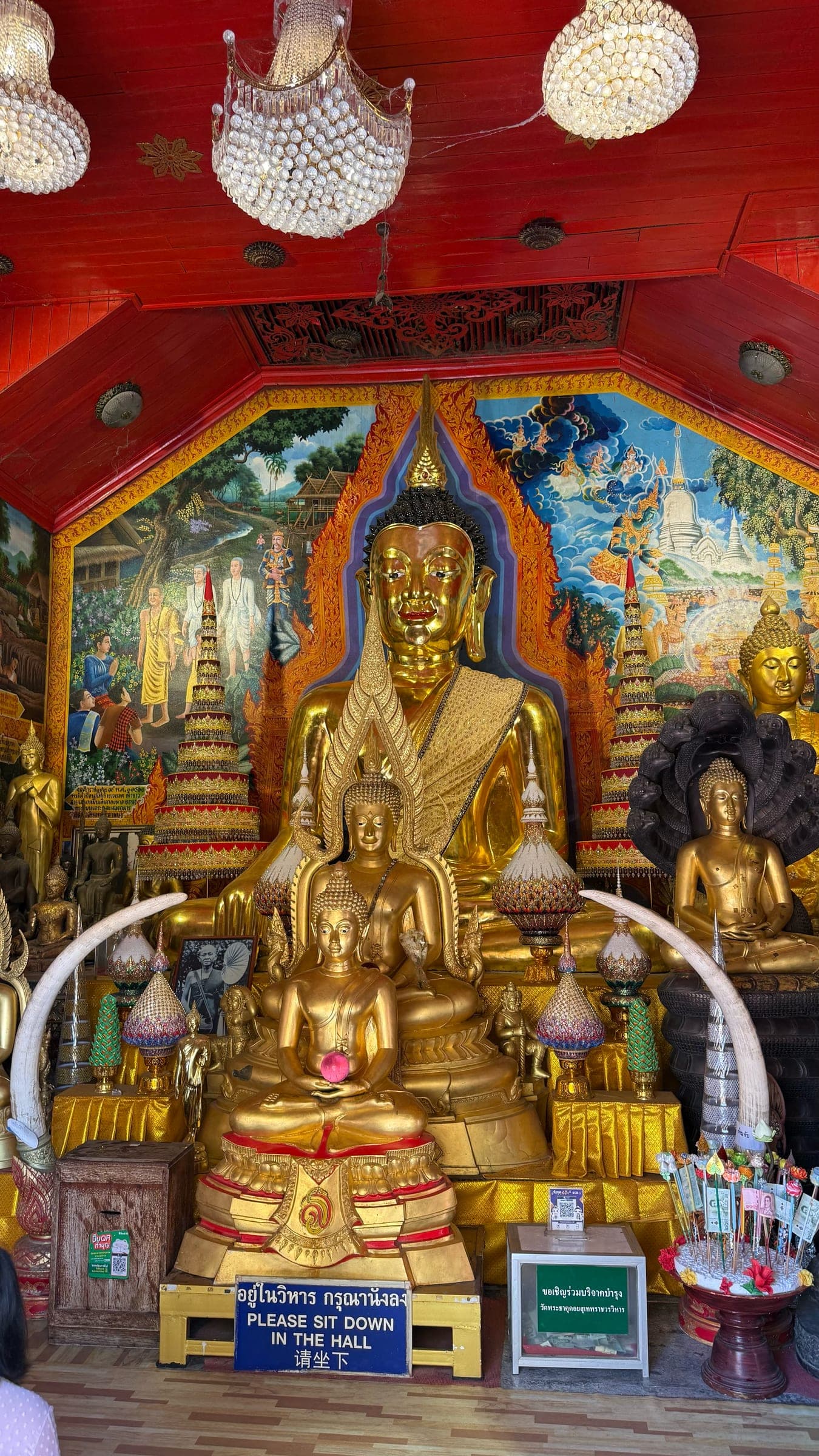 Gold seated Buddha statues, painted murals, and chandeliers inside a temple hall at Wat Phra That Doi Suthep
