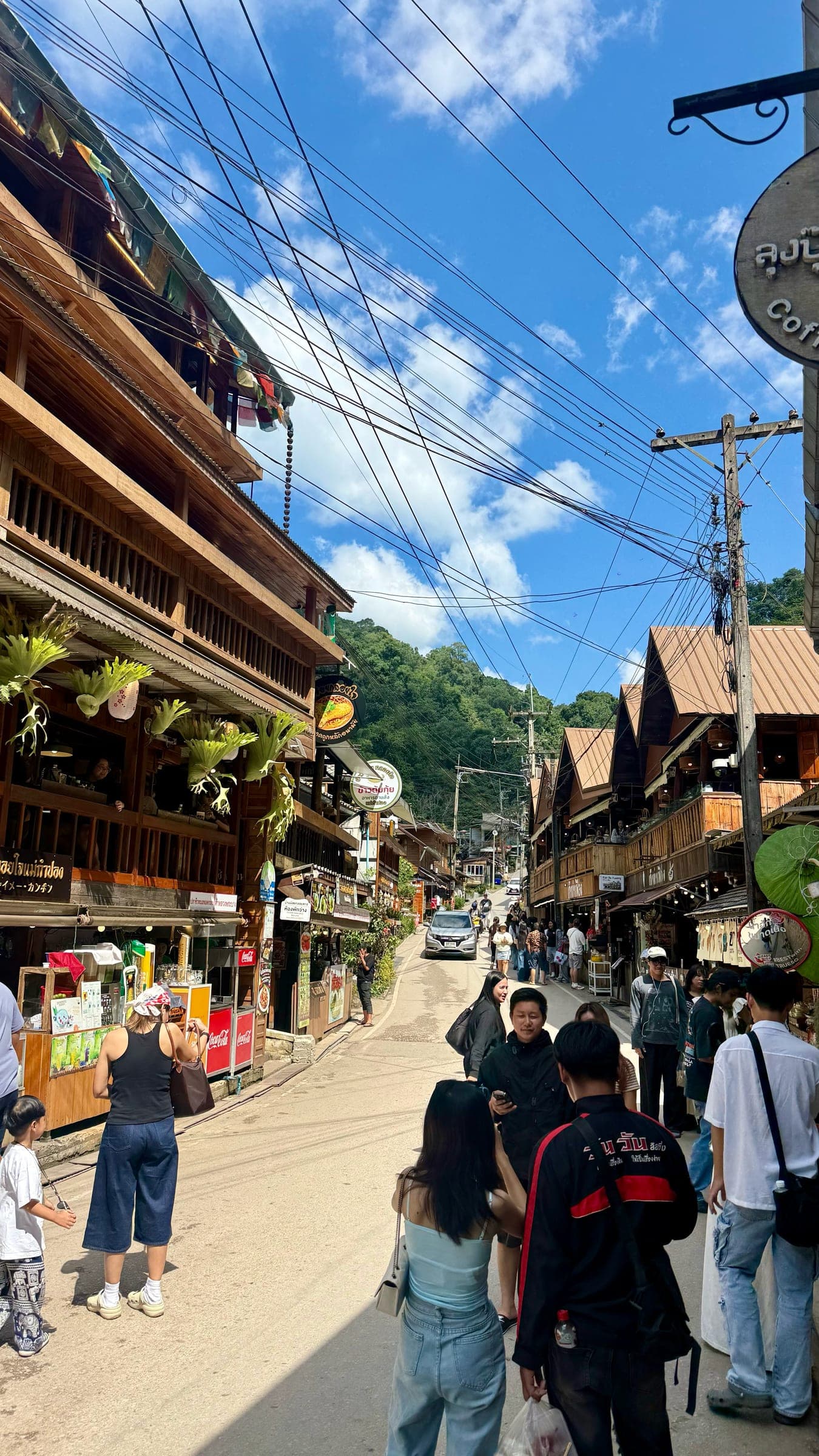 Visitors walking a sloping street lined with timber shophouses in the highland village of Mae Kampong, with forested hills behind