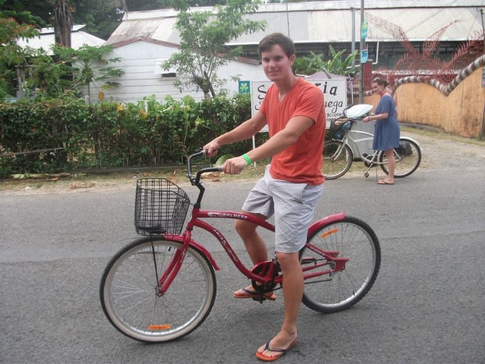 A traveler on a red beach cruiser bicycle with a front basket on a quiet town street, another cyclist behind