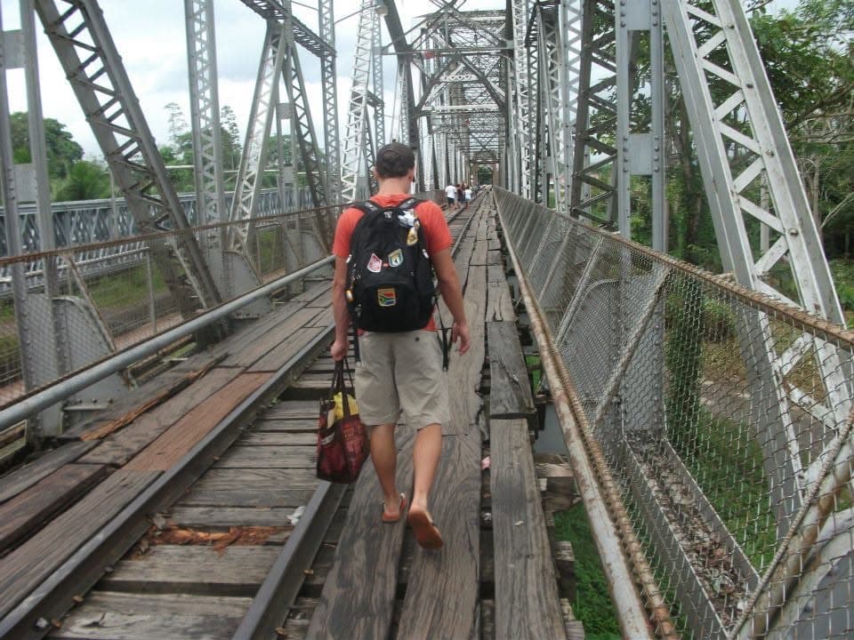 A backpacker walking on wooden planks laid over railway tracks across an old steel truss bridge at a border crossing