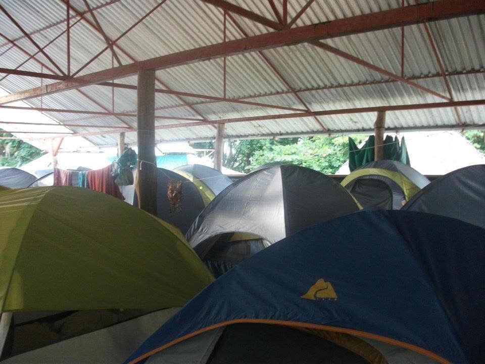 Rows of dome tents pitched under a corrugated metal roof at a budget backpacker campground, with laundry hanging on a line