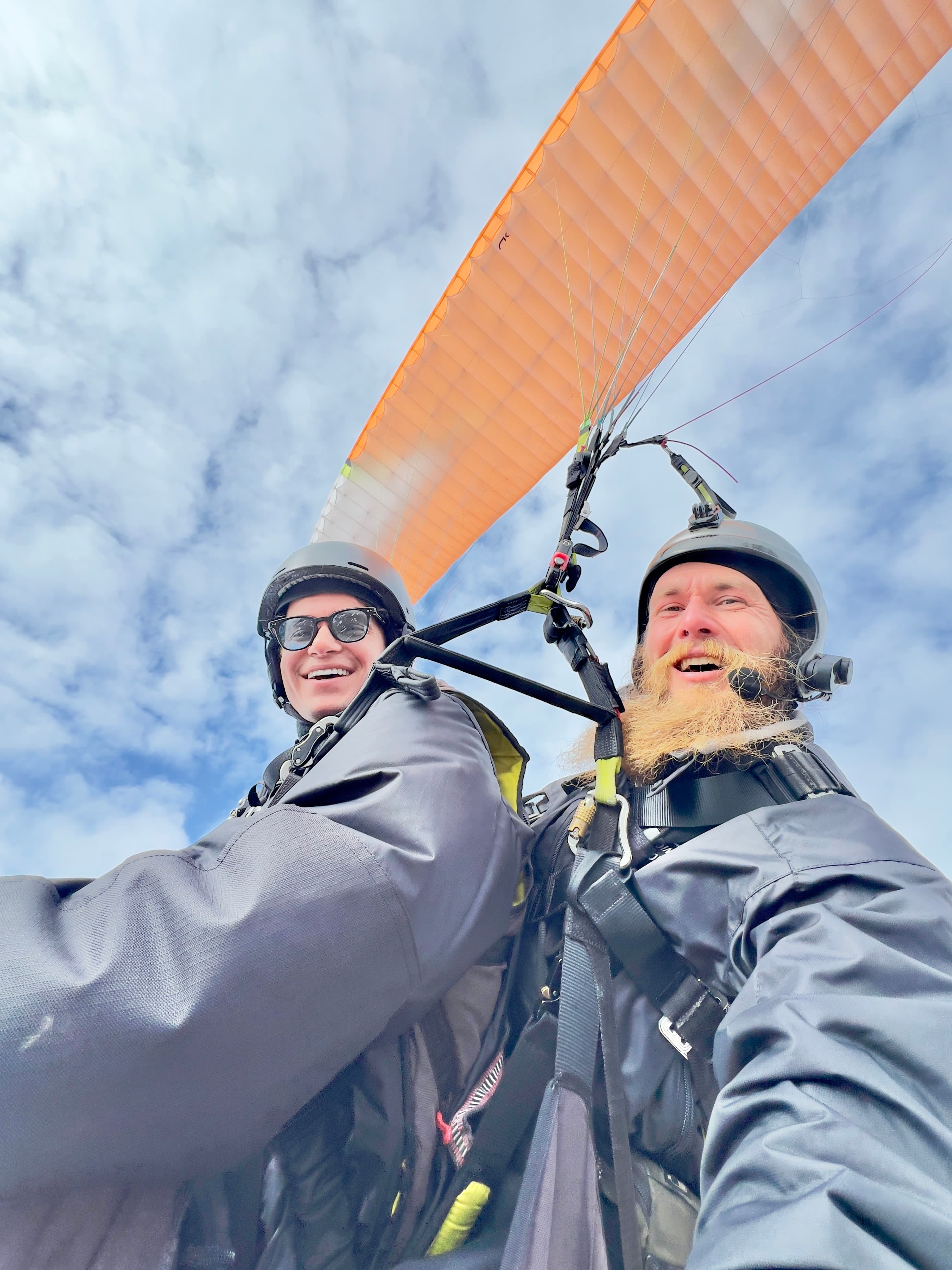 In-flight selfie of a traveler and a bearded tandem pilot smiling in harnesses beneath an orange paraglider wing high above the clouds over Gudauri