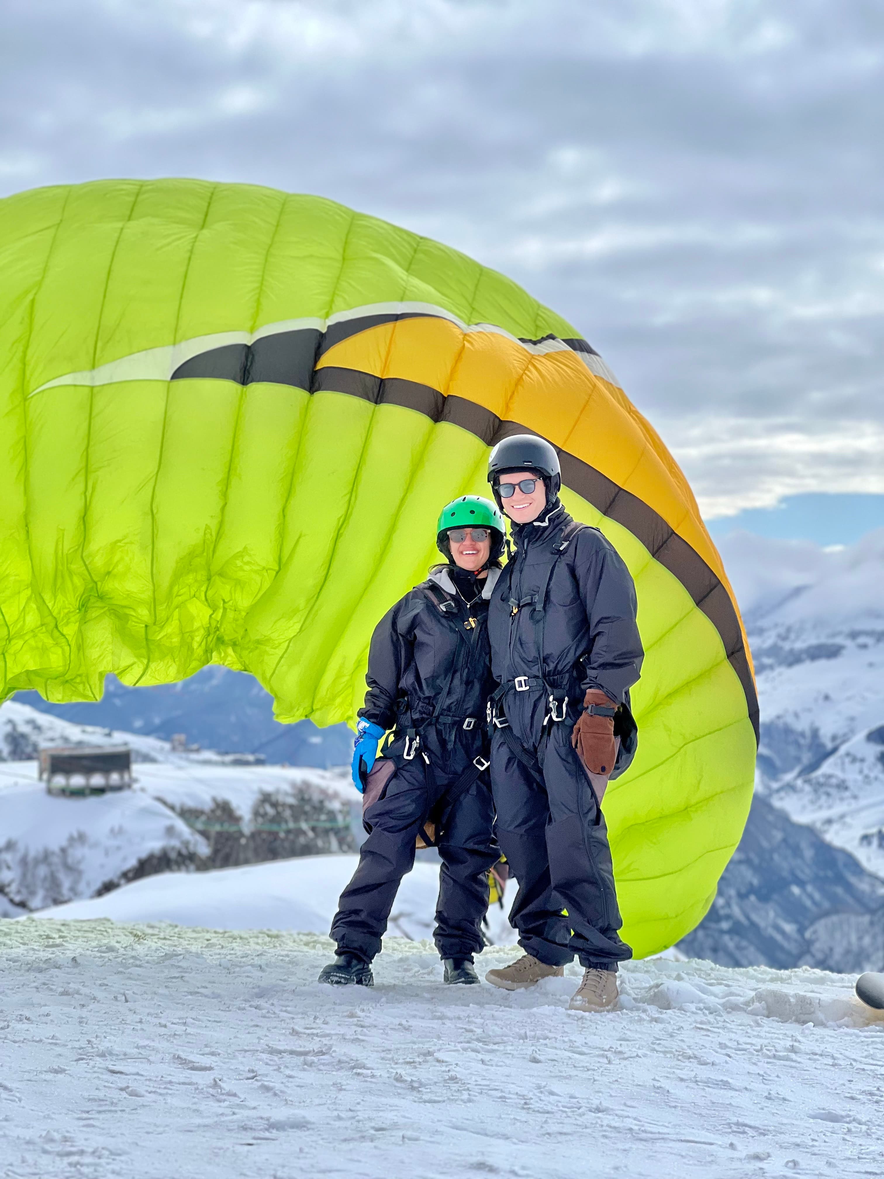 A couple in black flight suits and helmets standing in front of a bright green-and-yellow tandem paraglider canopy on the snow before takeoff at Gudauri