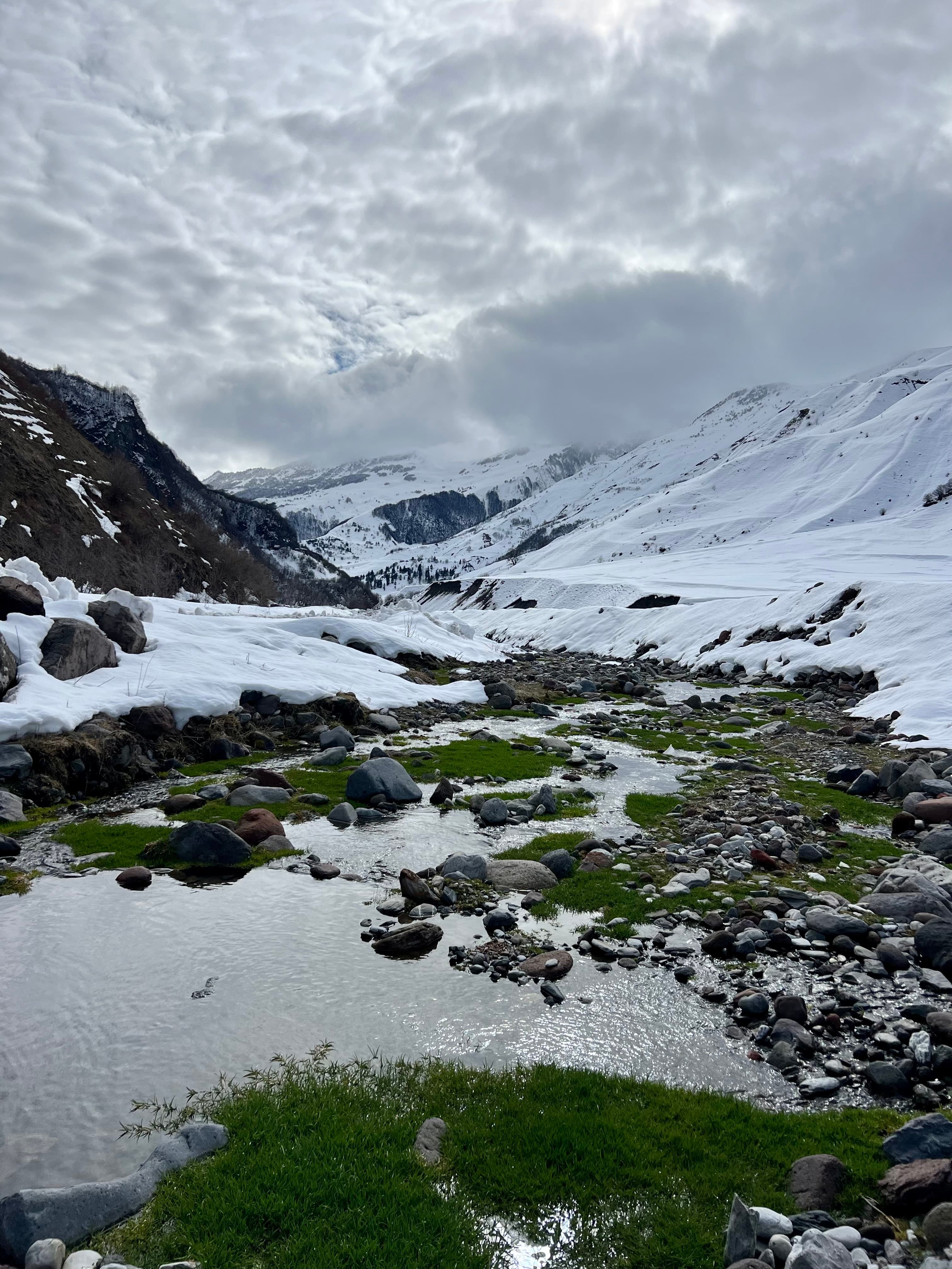A rocky snowmelt stream winding through a thawing valley of green grass and snow patches, framed by the snow-covered Greater Caucasus under a grey sky