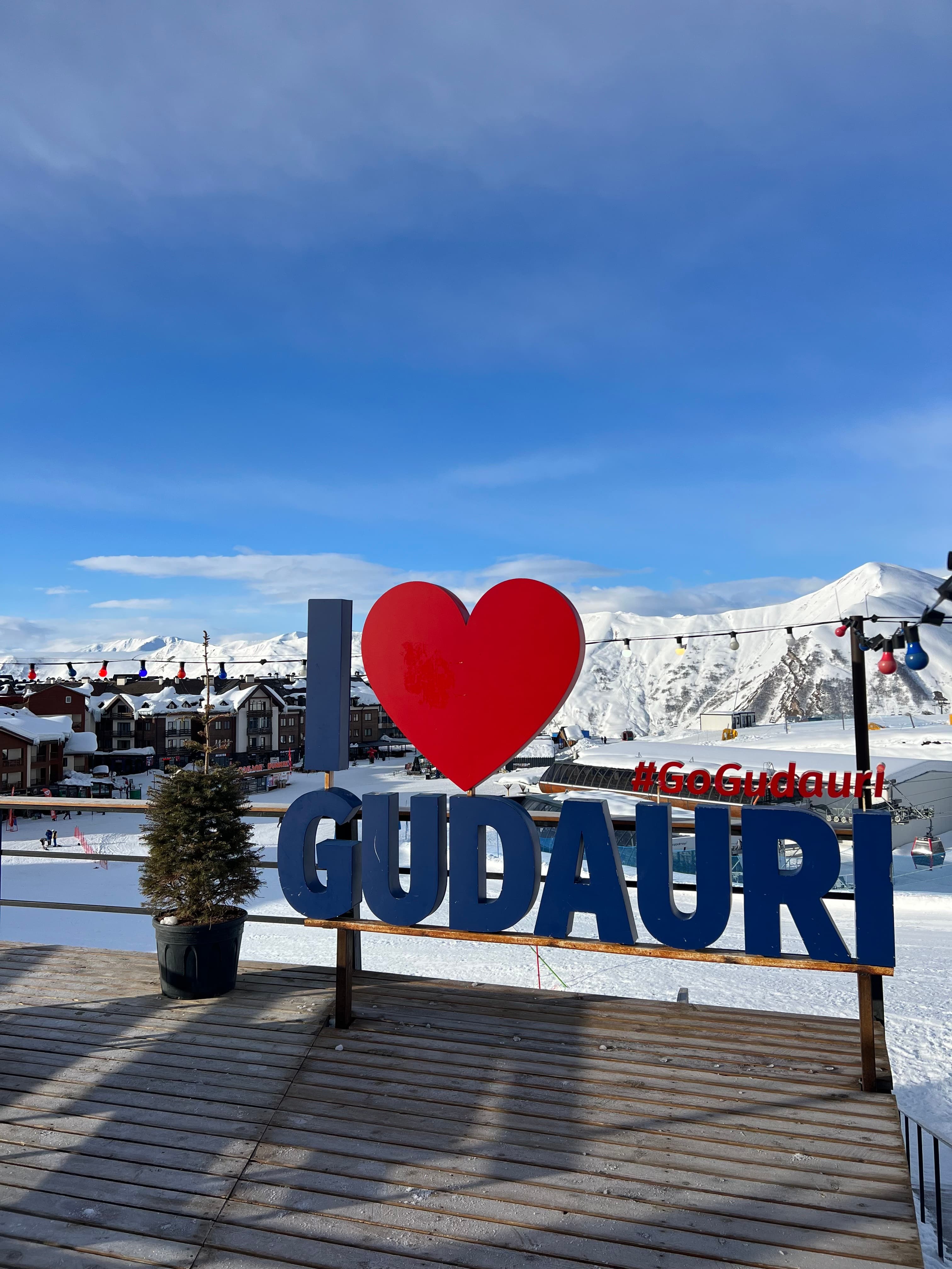 A red-and-blue 'I love Gudauri #GoGudauri' sign on a snowy deck above the ski village, gondola, and Caucasus mountains
