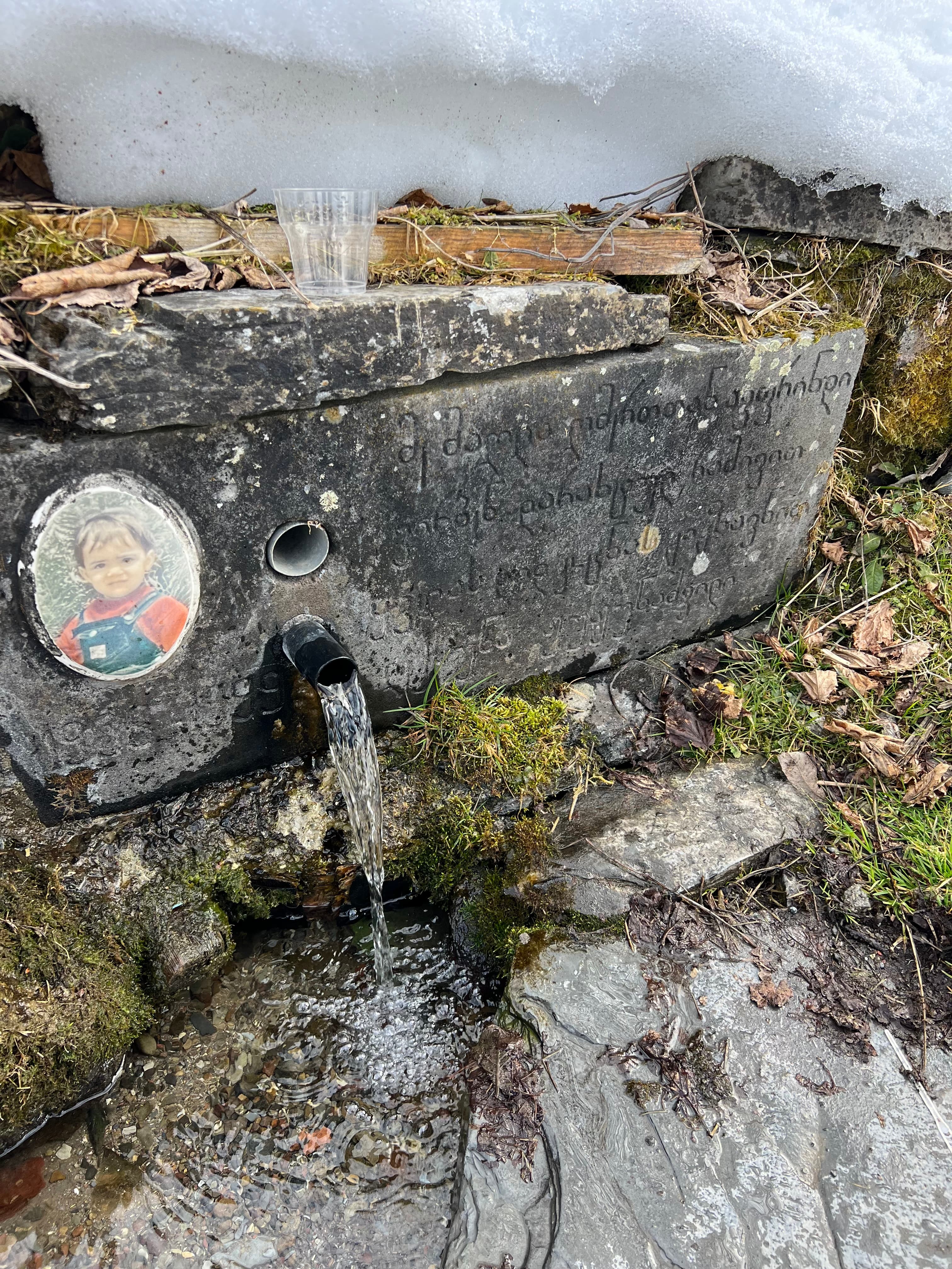 A roadside stone spring in the Caucasus, water running from a pipe below Georgian script carved into the slab and a child's photo memorial, with snow above