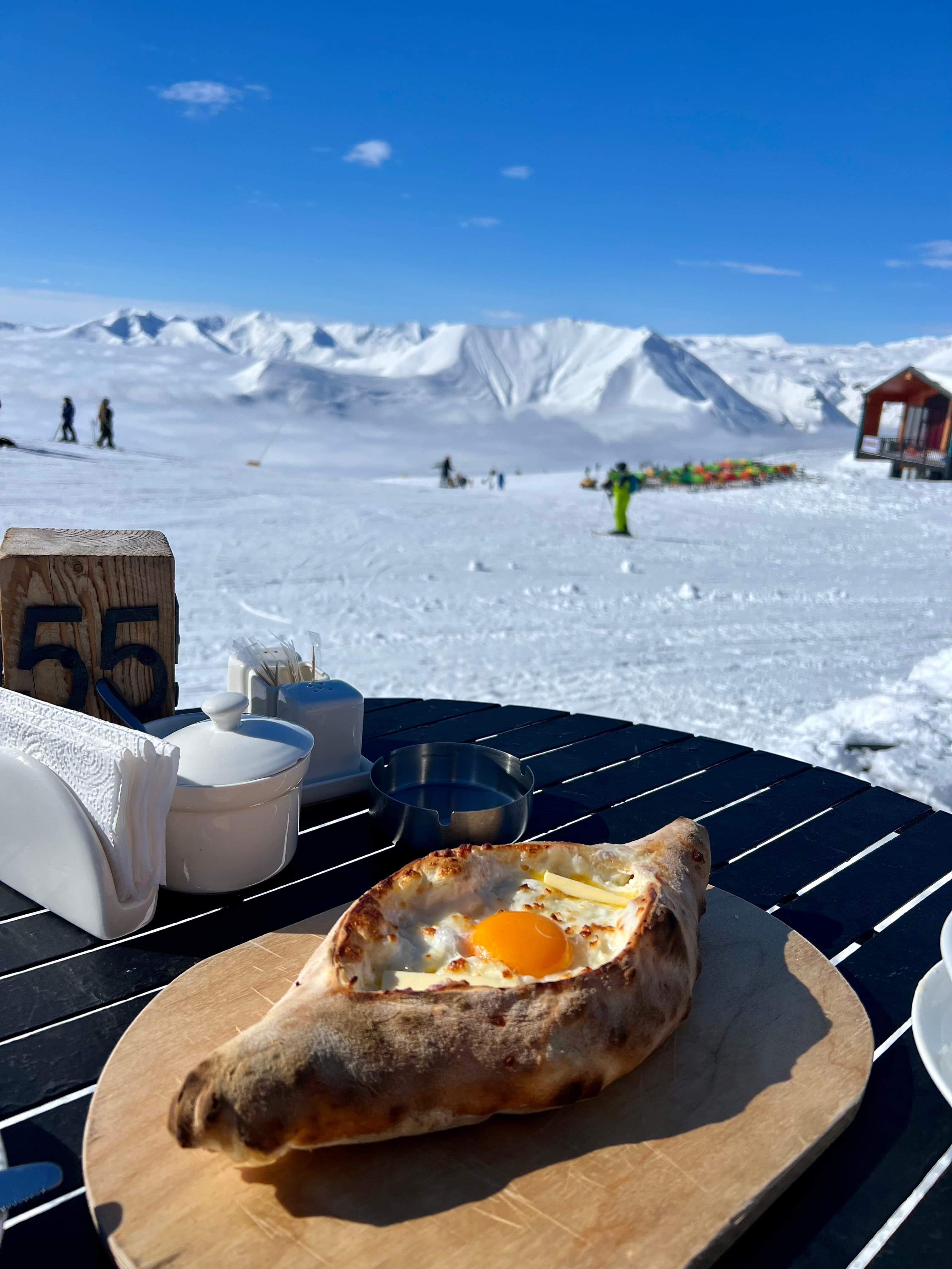 A boat-shaped Adjarian khachapuri with a runny egg yolk on a wooden board at a slopeside table, with skiers and the snow-covered Caucasus peaks behind, Gudauri