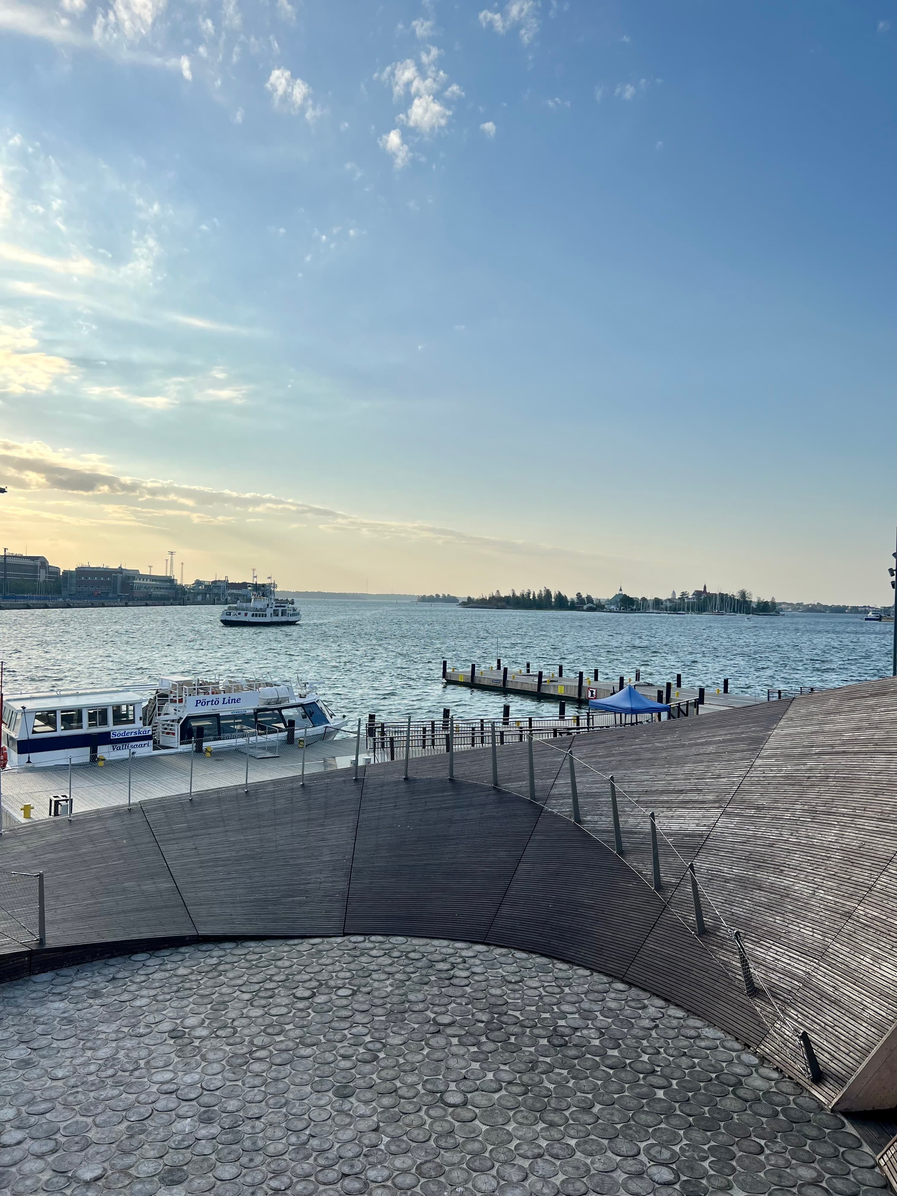 Small archipelago ferries at a pier on Helsinki's harbor at golden hour, seen from a curved wooden deck, with islands across the water