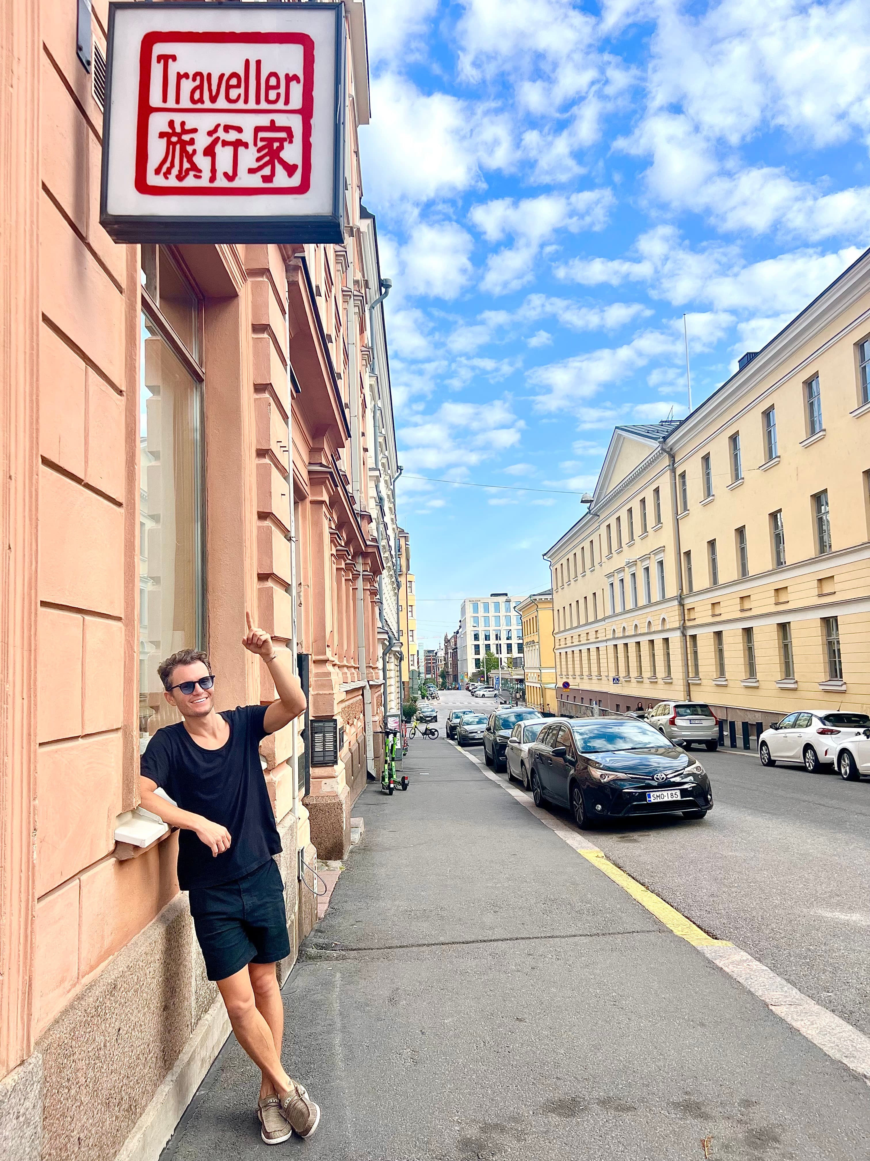 Standing beneath a 'Traveller' shop sign on a quiet sloping street of pastel neoclassical buildings in central Helsinki