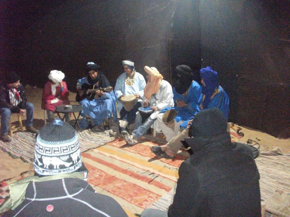Berber musicians playing guitar and hand drums for travelers seated on rugs inside a desert camp tent at night