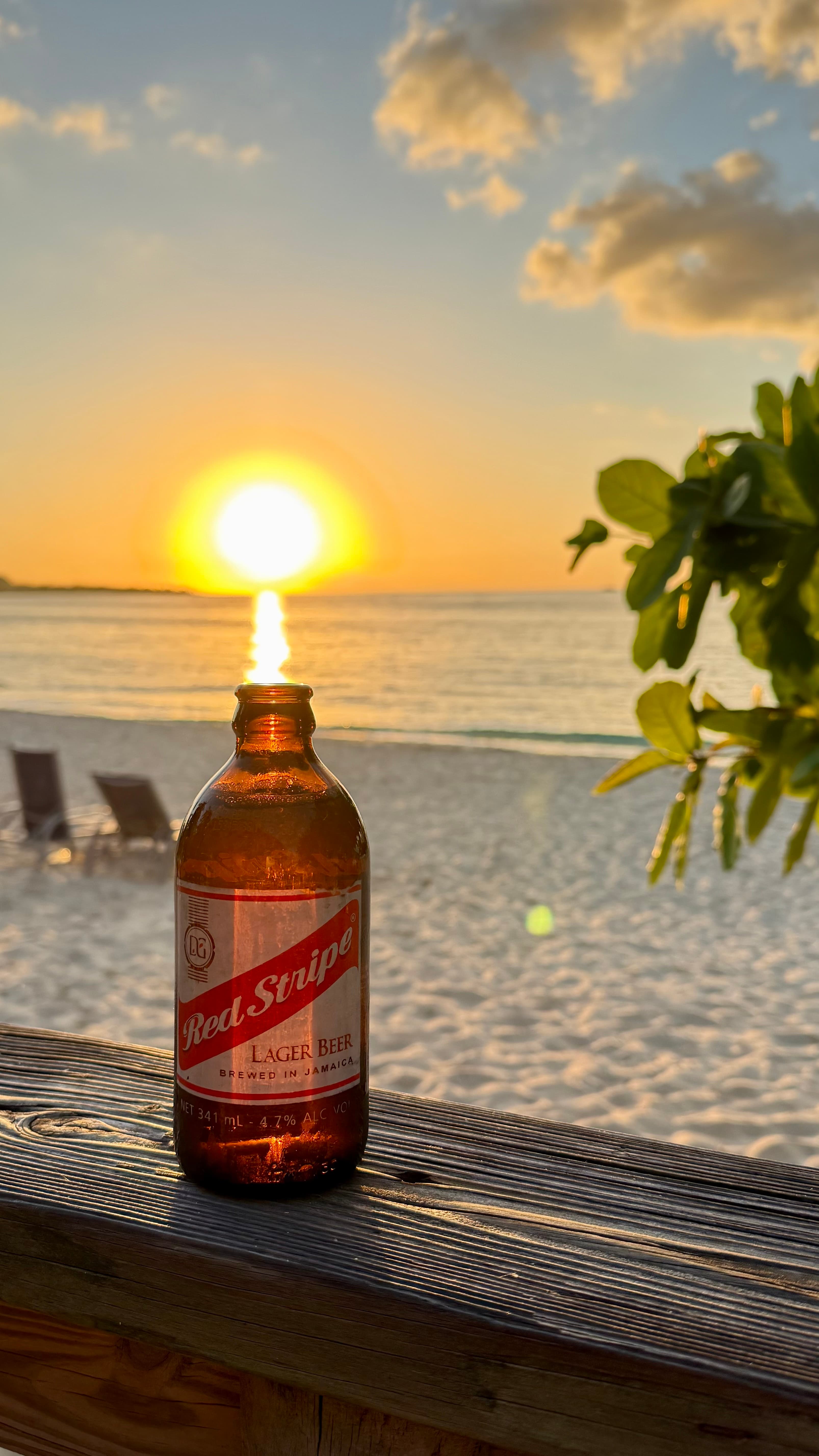 A Red Stripe lager bottle on a wooden rail with the sun setting over the white sand of Seven Mile Beach, Negril