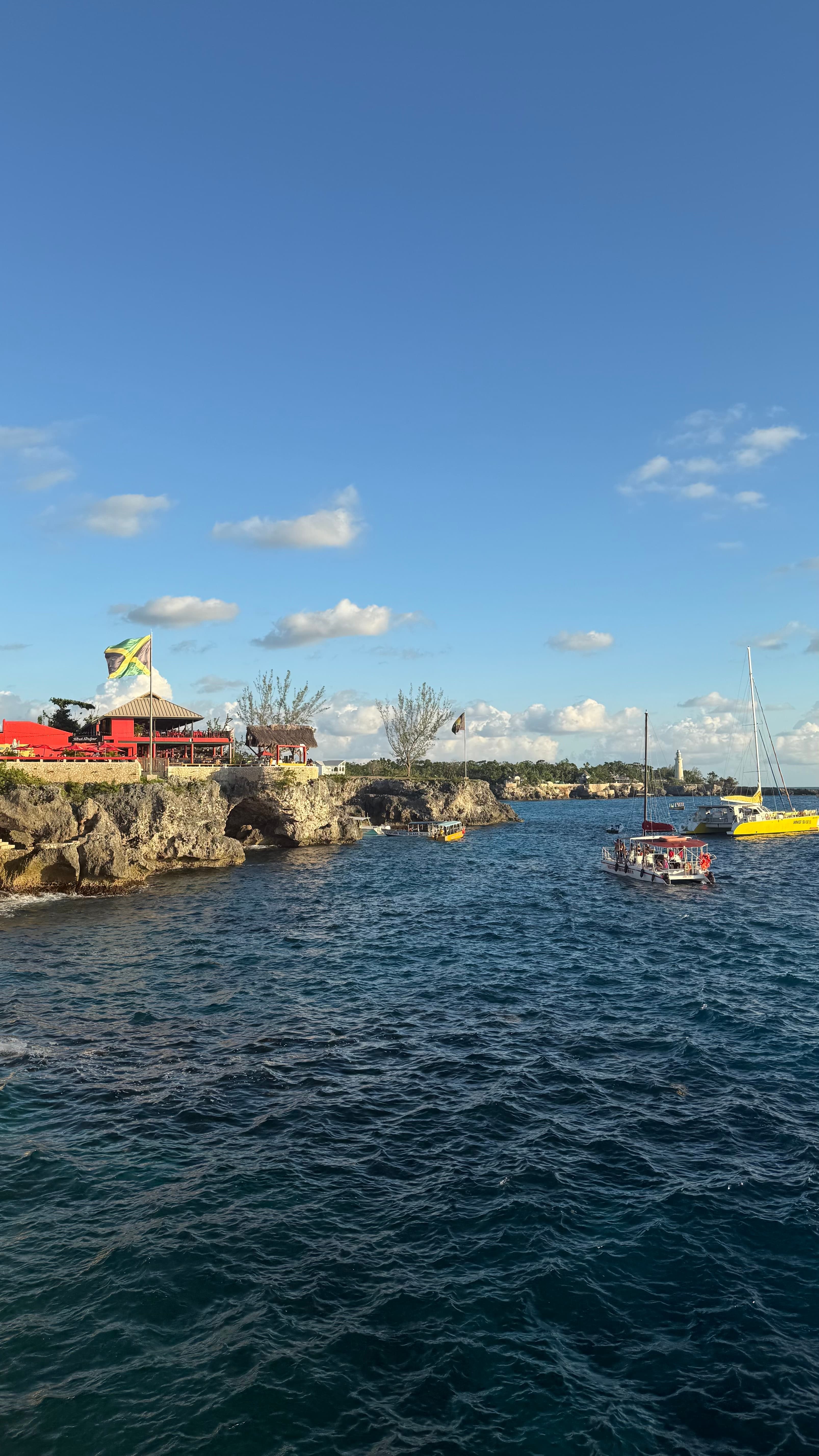 Rick's Cafe on the West End limestone cliffs flying a Jamaican flag above the water, with boats and the Negril Lighthouse in the distance