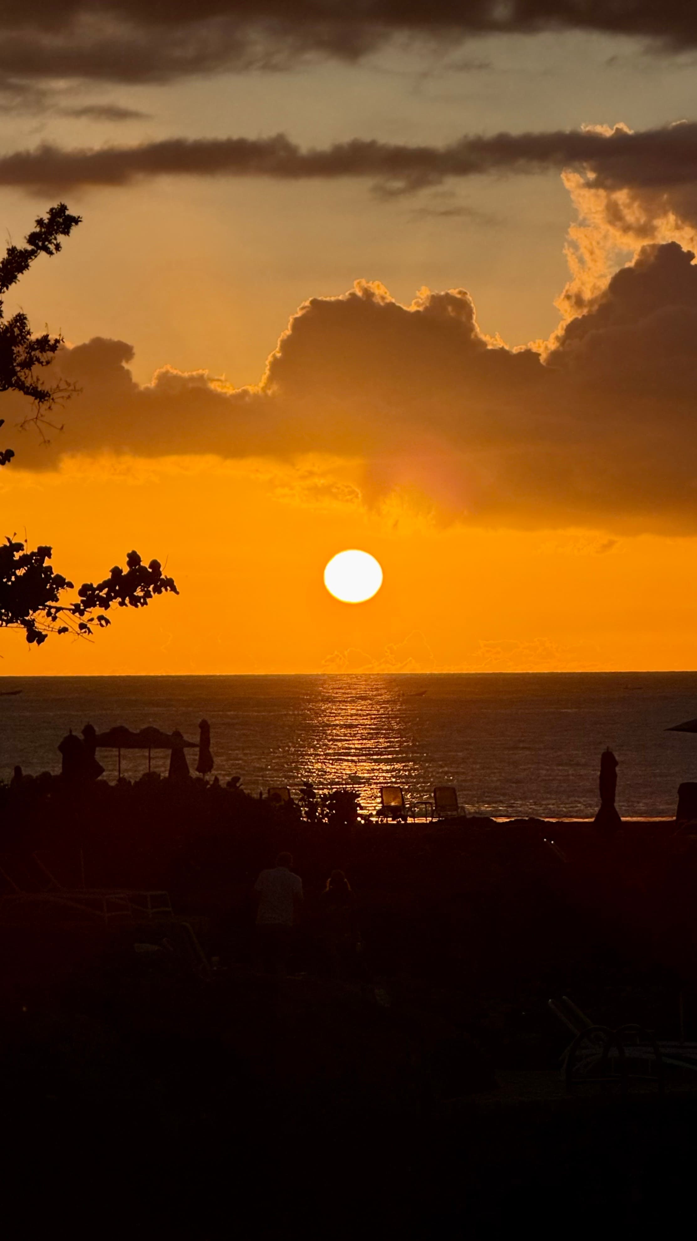 The sun setting into the sea beyond silhouetted trees and beach loungers on Negril's west-facing coast, Jamaica