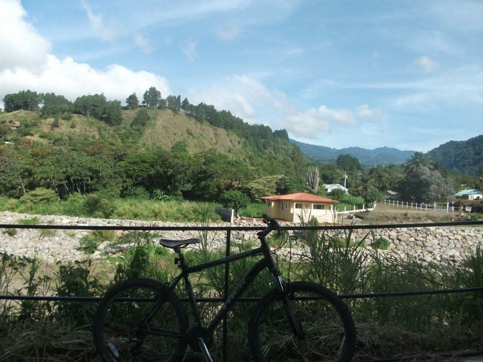 A black mountain bike leaning on a railing above a rocky riverbed, with a small orange-roofed house and pine-covered hills beyond, Boquete