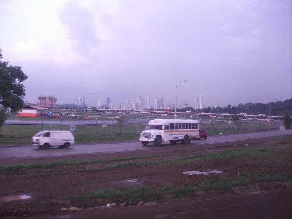 A white school-bus-style local bus on a highway under a stormy sky, with the Panama City skyline on the horizon