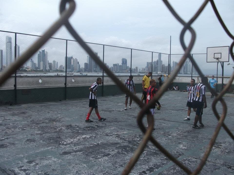 Young players in striped jerseys on a fenced concrete soccer court, seen through chain-link, with the Panama City skyline across the bay