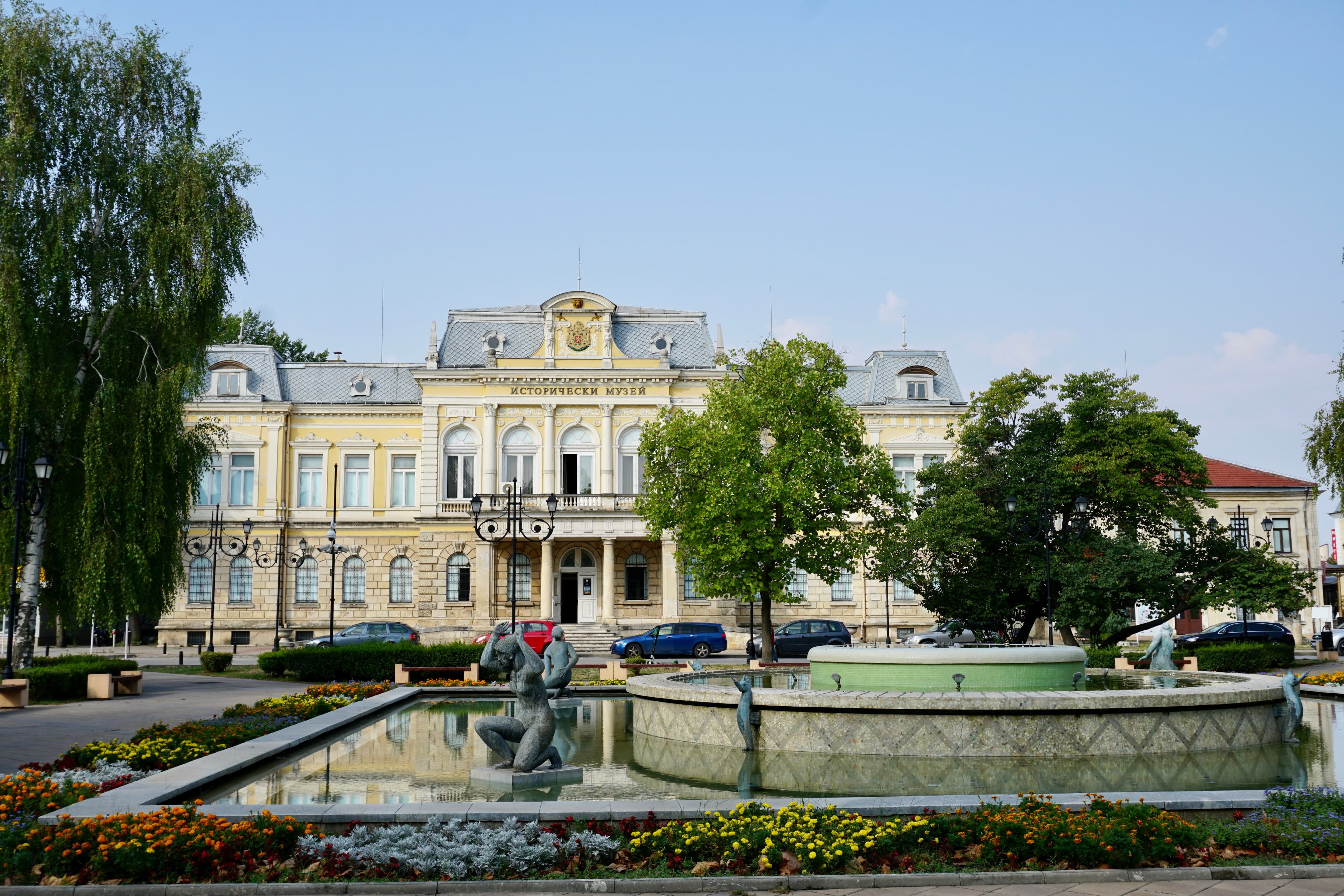 The yellow neo-baroque Regional Historical Museum behind a fountain and flower beds in Ruse