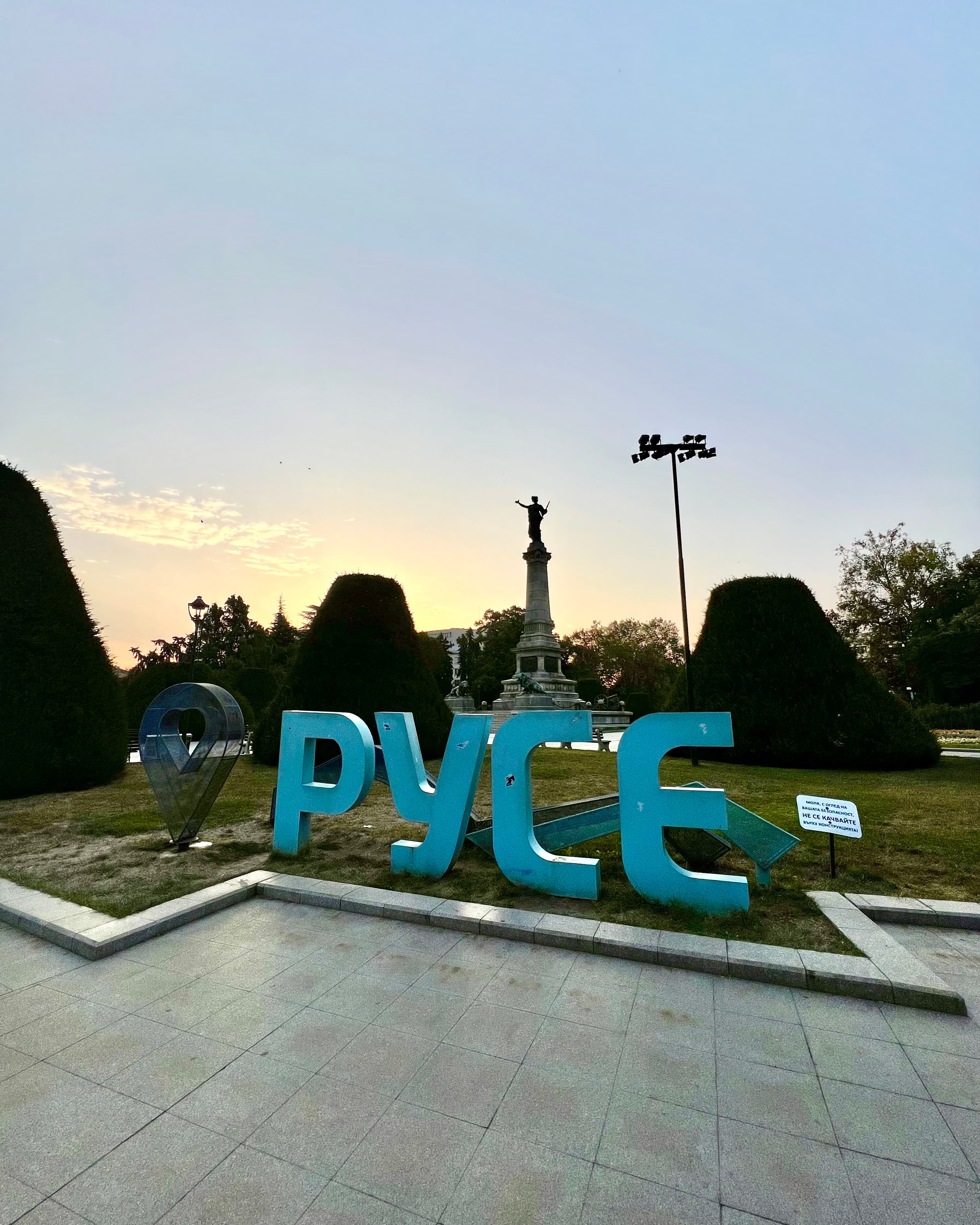 Large blue Cyrillic letters spelling Ruse in a park at dusk, with the Monument of Liberty rising on its column behind