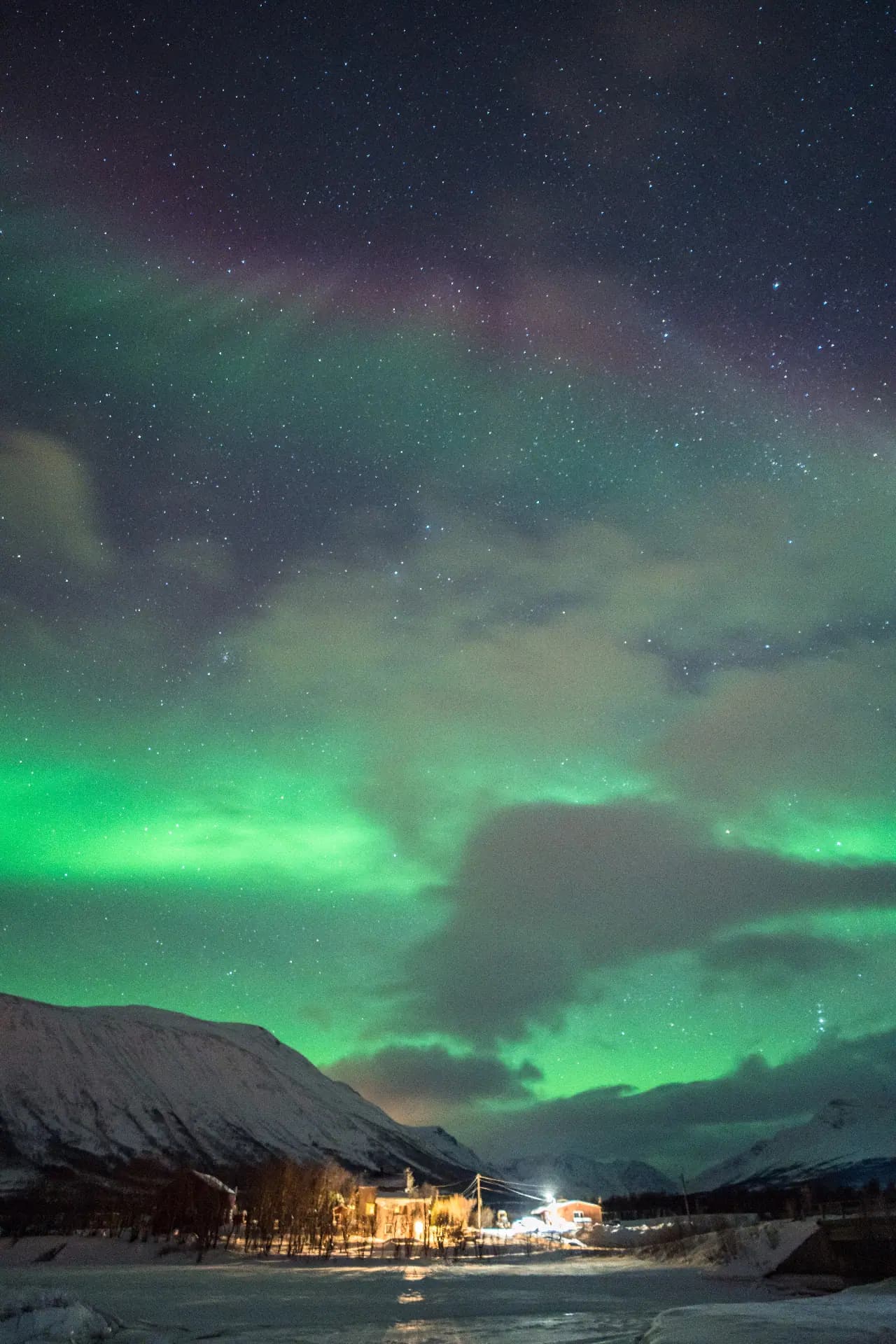 Green aurora glowing above clouds and a riverside village outside Tromsø